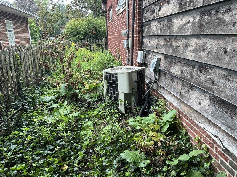 Conducted estimate for complete HVAC system replacement at residential property. Evaluated existing outdoor condenser unit located on side of building, which is surrounded by overgrown vegetation and shows signs of aging infrastructure including oxidized copper refrigerant lines and weathered electrical conduit. Documented current system condition with photographs for insurance claim purposes, noting the unit's accessibility issues due to dense plant growth and the deteriorated state of associated piping and utility connections at the building's foundation.