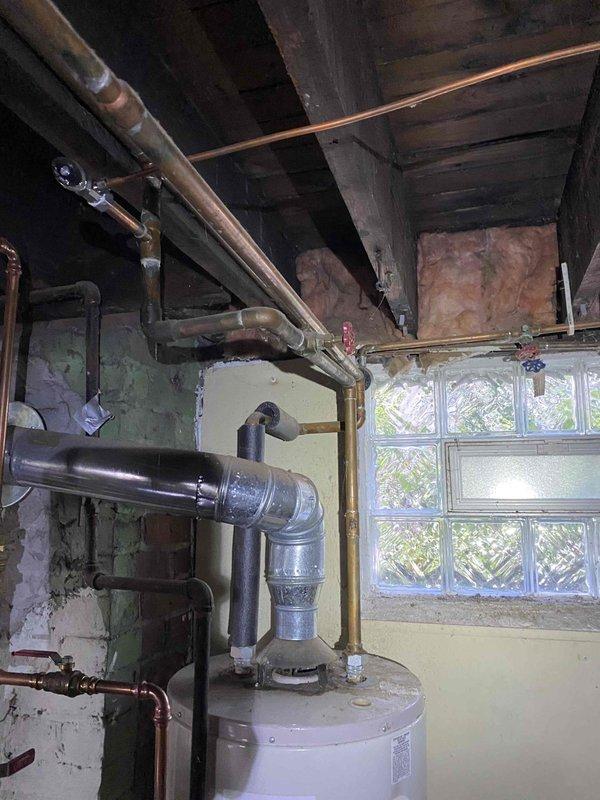 Responded to service call for water accumulation around boiler system. Upon inspection, identified water heater in basement utility area with deteriorated plumbing connections and visible moisture issues. Examined copper piping, galvanized vent stack, and associated mechanical components for signs of leakage. Documented conditions including aged infrastructure, corroded piping with dark deterioration along vertical runs, and wall staining indicative of previous water exposure. Assessed water meter connections, pressure relief valve, and all visible joints for active leaks or seepage points to determine source of reported water accumulation.