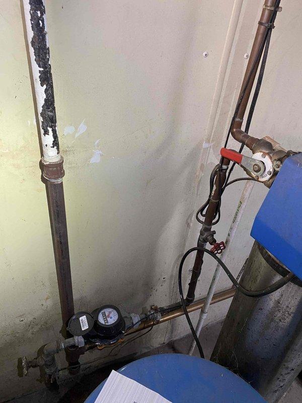 Responded to service call for water accumulation around boiler system. Upon inspection, identified water heater in basement utility area with deteriorated plumbing connections and visible moisture issues. Examined copper piping, galvanized vent stack, and associated mechanical components for signs of leakage. Documented conditions including aged infrastructure, corroded piping with dark deterioration along vertical runs, and wall staining indicative of previous water exposure. Assessed water meter connections, pressure relief valve, and all visible joints for active leaks or seepage points to determine source of reported water accumulation.