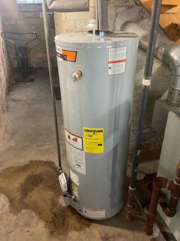 Responded to service call for leaking water heater under home protection plan membership. Upon arrival, inspected 8-12 year old tanked water heater unit located in basement utility area. Visual assessment revealed active leak originating from lower copper pipe connections with yellow brass fittings. Observed significant mineral deposits and white residue buildup around pipe connection points at base of unit, indicating chronic moisture exposure and potential fitting deterioration. Documented condition of water heater showing typical age-related wear, corrosion at connection points, and moisture staining on concrete floor surrounding unit base. Photographed unit specifications, safety labels, plumbing connections, and leak evidence for service records. Water heater displays standard residential installation with proper venting and utility connections in unfinished basement environment.