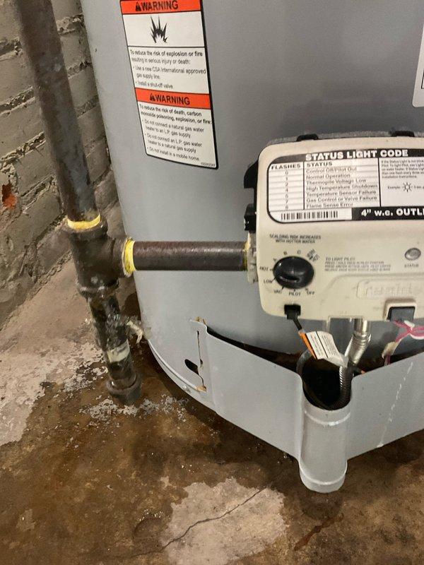 Responded to service call for leaking water heater under home protection plan membership. Upon arrival, inspected 8-12 year old tanked water heater unit located in basement utility area. Visual assessment revealed active leak originating from lower copper pipe connections with yellow brass fittings. Observed significant mineral deposits and white residue buildup around pipe connection points at base of unit, indicating chronic moisture exposure and potential fitting deterioration. Documented condition of water heater showing typical age-related wear, corrosion at connection points, and moisture staining on concrete floor surrounding unit base. Photographed unit specifications, safety labels, plumbing connections, and leak evidence for service records. Water heater displays standard residential installation with proper venting and utility connections in unfinished basement environment.