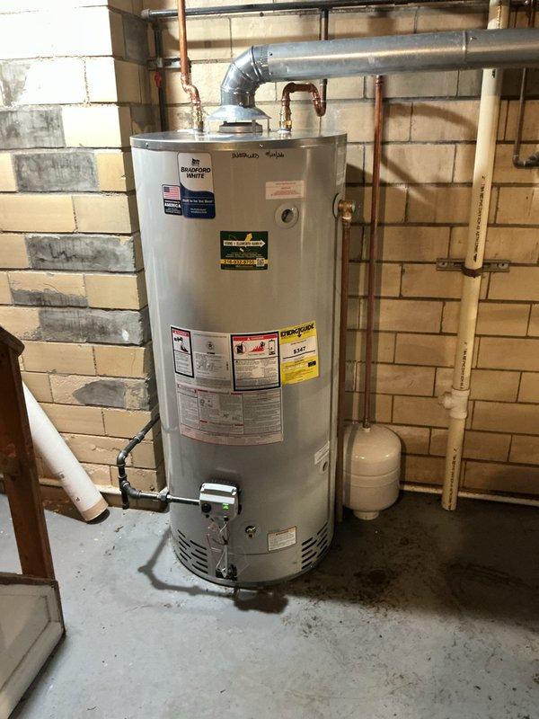 Responded to service call for malfunctioning water heater in residential basement utility area. Upon inspection, determined that the existing gas-fired water heater tank had failed and required replacement. Removed old unit and installed new white cylindrical residential gas water heater with proper venting through metallic flue pipe. Connected copper water supply lines to inlet and outlet ports on new tank. Verified proper installation of expansion tank mounted on adjacent wall. Tested all connections for leaks and confirmed proper operation of new water heater system.