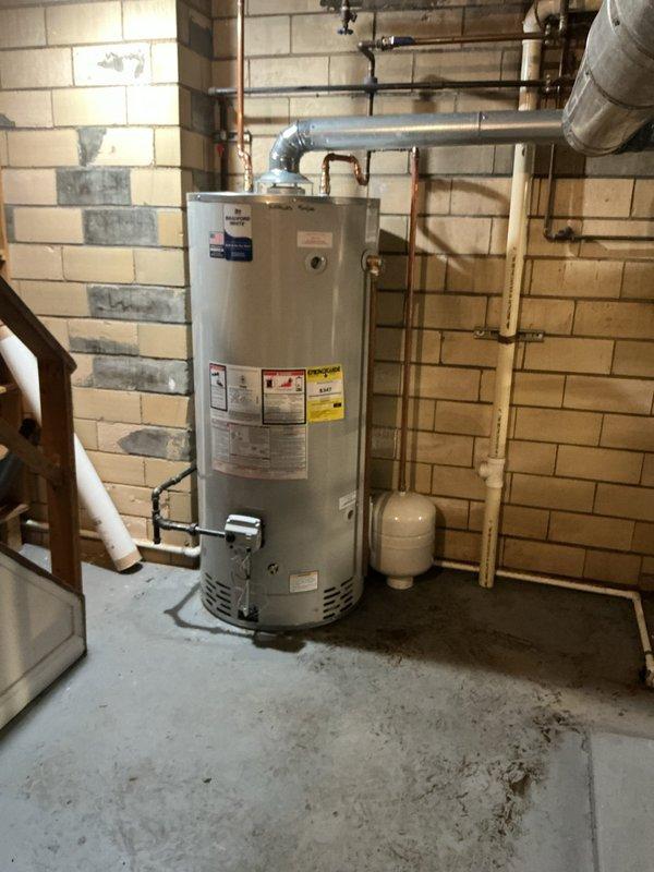 Responded to service call for malfunctioning water heater in residential basement utility area. Upon inspection, determined that the existing gas-fired water heater tank had failed and required replacement. Removed old unit and installed new white cylindrical residential gas water heater with proper venting through metallic flue pipe. Connected copper water supply lines to inlet and outlet ports on new tank. Verified proper installation of expansion tank mounted on adjacent wall. Tested all connections for leaks and confirmed proper operation of new water heater system.
