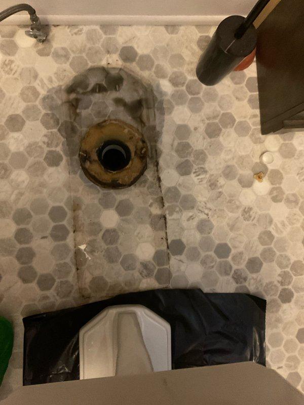 Responded to a water leak at the laundry drain pipe during normal business hours. Upon inspection, identified significant corrosion and rust buildup on the drain fixture assembly, with evidence of long-term moisture exposure and mineral deposits causing deterioration of the drain components. Documented the condition of the hexagonal tile floor surrounding the drain, noting water staining, grime accumulation in grout lines, and visible wear patterns consistent with chronic water leakage. The drain fixture showed advanced oxidation on metal components with rust-colored staining extending to surrounding floor surfaces. Photographed the affected areas for service records, capturing the extent of corrosion on the concentric ring assembly and the deteriorated condition of the floor tiles adjacent to the leak source.
