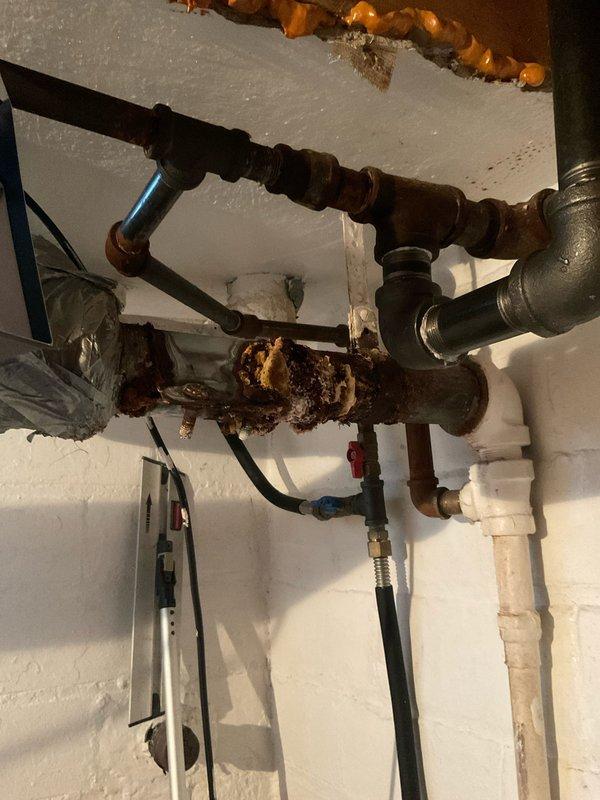 Responded to service call for toilet leak from upstairs bathroom causing water damage to ceiling below. Upon inspection, identified severely corroded horizontal drain pipe in basement utility area with extensive rust deterioration and compromised structural integrity. The corroded pipe section was causing water to leak through deteriorated areas when upstairs toilet was flushed, resulting in water infiltration into the ceiling below. Documented existing plumbing infrastructure including multiple connection points, PVC piping, and associated fittings. Photographed corroded pipe section showing heavy rust accumulation and flaking metal, as well as upstairs bathroom with white porcelain toilet and decorative tile work to document service location. Recommended immediate replacement of corroded drain pipe section to prevent further water damage and restore proper drainage function.