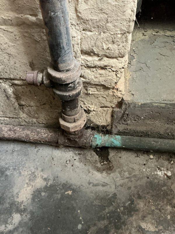 Responded to service call for 15-year-old boiler in basement exhibiting loud rumbling sound followed by perking noise. Upon arrival, performed visual inspection of boiler system and associated plumbing components, documenting aged galvanized piping with visible corrosion and deteriorated fittings. Examined copper piping connections, pressure vessel, and control valves for signs of kettling, sediment buildup, or mechanical failure that could cause reported noise issues. Photographed system components including corroded horizontal pipe sections showing green patina oxidation and worn threaded unions near floor level. Assessed overall condition of 12-13 year old heating system and surrounding infrastructure showing significant age-related wear.