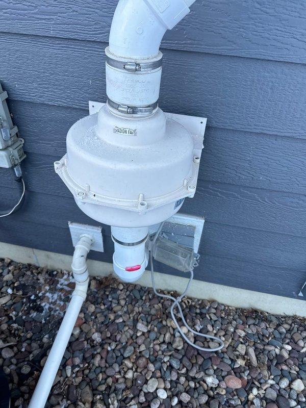 Responded to customer call regarding noise from mechanical room. Diagnosed failed radon mitigation fan due to ice buildup. System includes a RadonAway GP501 fan unit mounted to exterior wall with PVC piping connections. Provided customer with replacement fan estimate.