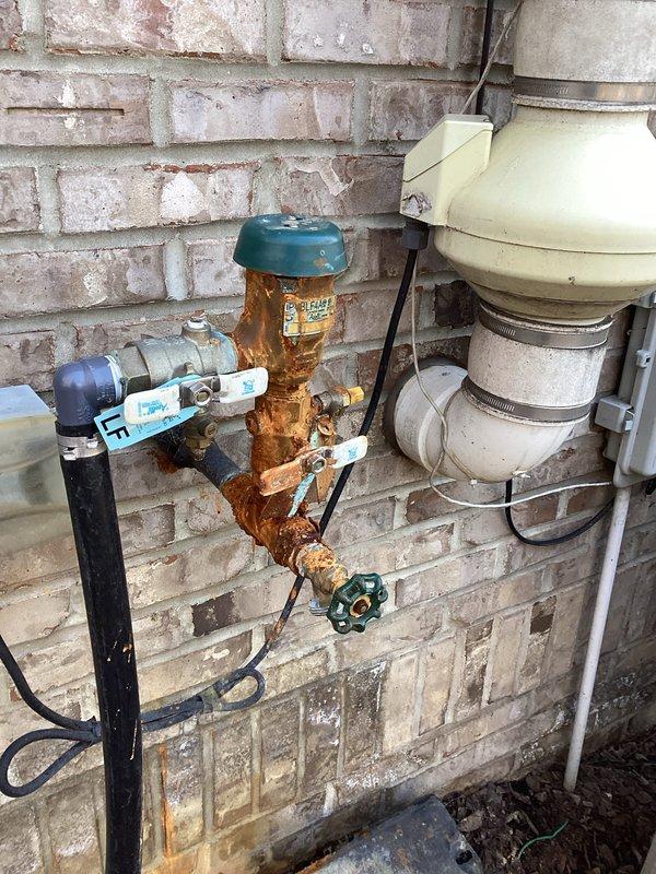 Inspected severely corroded backflow preventer for irrigation system mounted on exterior brick wall. Documented extensive rust damage and deterioration on copper pipe assembly with green valve handle. Provided customer with parts estimate for backflow preventer replacement; parts on order with expected availability within 10 days.