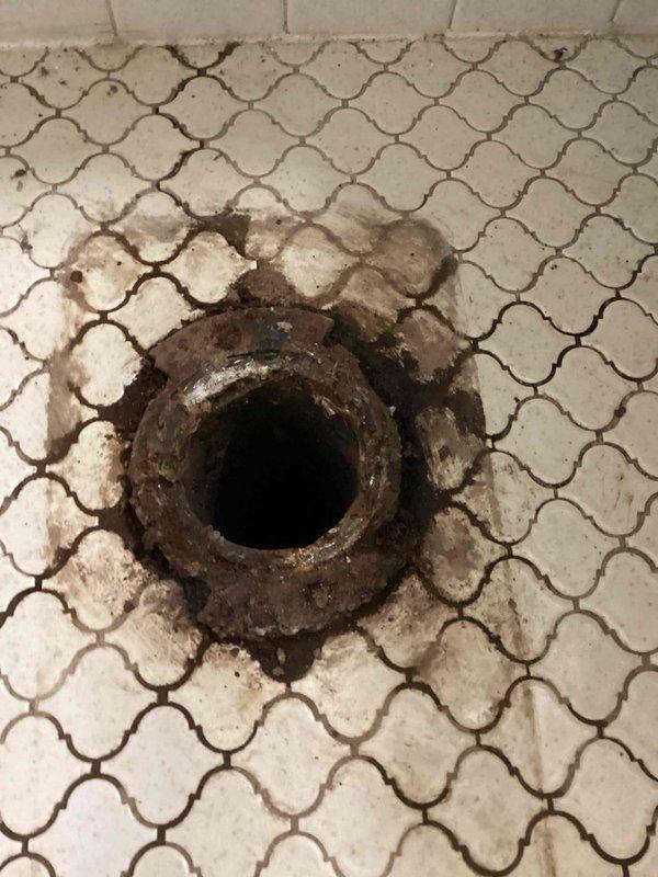 Responded to service call for toilet main pipe replacement. Upon inspection, discovered severely corroded and deteriorated toilet flange with visible rust and mineral deposits. Identified cast iron drain line experiencing internal leaking from upstairs bathroom into basement ceiling below. Documented extensive corrosion on floor drain and compromised drainage system. Due to inaccessible nature of leak and extent of deterioration in cast iron piping system, recommended complete repipe of toilet flange and conversion of cast iron drain line to PVC. Captured photographic documentation showing corroded drain components, water damage staining on surrounding tile surfaces, and exposed basement infrastructure including deteriorated cast iron vertical stack and overhead plumbing lines. Scheduled follow-up assessment for comprehensive quote on toilet flange replacement and full drain line conversion project.