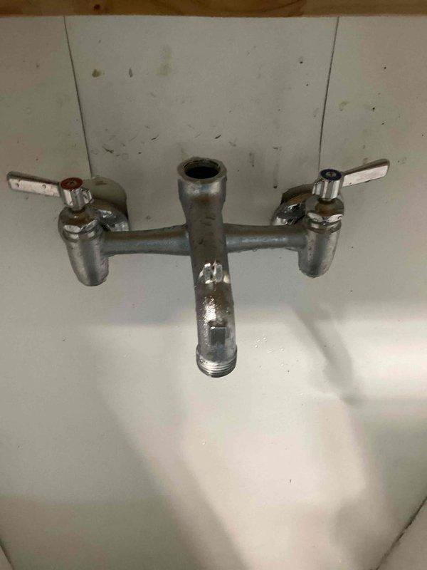 Responded to service call for leaking wall-mounted utility faucet in garage. Upon inspection, discovered the original center plug fitting between the hot and cold handles was missing and had been temporarily replaced with an incorrect-sized 3/4" PVC plug that did not properly seal. Measured the threaded opening and determined the correct replacement part requires a 7/8" threaded plug. The aging chrome-plated fixture showed significant wear, corrosion, and deterioration of the finish. Documented the faucet model and configuration with photographs for parts identification and ordering purposes, as the specific manufacturer and model information was not readily visible on the fixture. Advised that further research would be conducted to identify the faucet brand and source the proper OEM replacement plug fitting to restore the unit to proper working condition and eliminate the leak.