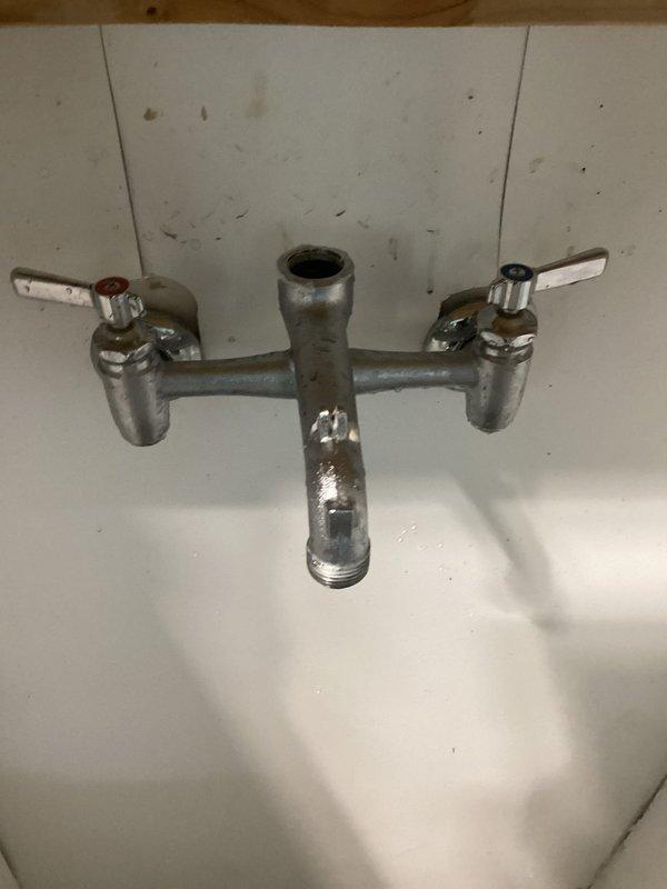 Responded to service call for leaking wall-mounted utility faucet in garage. Upon inspection, discovered the original center plug fitting between the hot and cold handles was missing and had been temporarily replaced with an incorrect-sized 3/4" PVC plug that did not properly seal. Measured the threaded opening and determined the correct replacement part requires a 7/8" threaded plug. The aging chrome-plated fixture showed significant wear, corrosion, and deterioration of the finish. Documented the faucet model and configuration with photographs for parts identification and ordering purposes, as the specific manufacturer and model information was not readily visible on the fixture. Advised that further research would be conducted to identify the faucet brand and source the proper OEM replacement plug fitting to restore the unit to proper working condition and eliminate the leak.