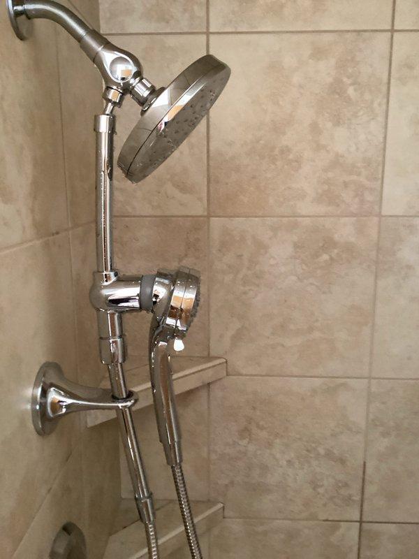 Removed existing shower head fixture and installed customer-supplied dual shower system featuring a rainfall-style circular shower head mounted on an adjustable curved chrome arm and a handheld sprayer with flexible metal hose and wall-mounted bracket. All components were properly secured to the tiled wall, connections were tested for leaks, and the installation was verified for proper water flow and functionality.