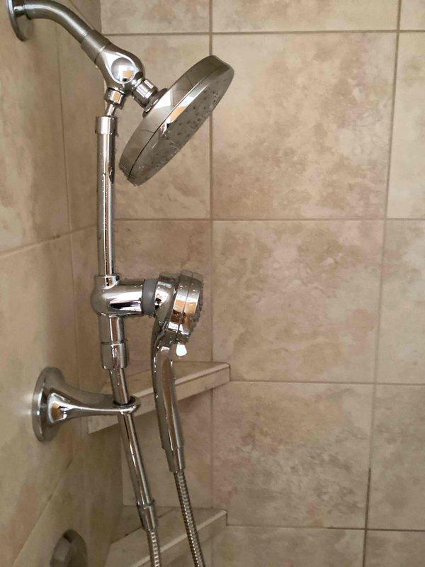 Removed existing shower head fixture and installed customer-supplied dual shower system featuring a rainfall-style circular shower head mounted on an adjustable curved chrome arm and a handheld sprayer with flexible metal hose and wall-mounted bracket. All components were properly secured to the tiled wall, connections were tested for leaks, and the installation was verified for proper water flow and functionality.