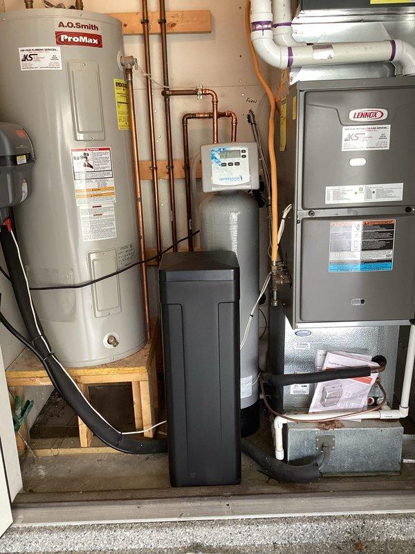 Completed installation of two-piece water softener system with custom-ordered smaller capacity brine tank to accommodate space constraints in residential utility area. Unit was professionally integrated with existing plumbing infrastructure using copper piping connections, positioned centrally between water heater and HVAC furnace, with digital control panel mounted and operational.