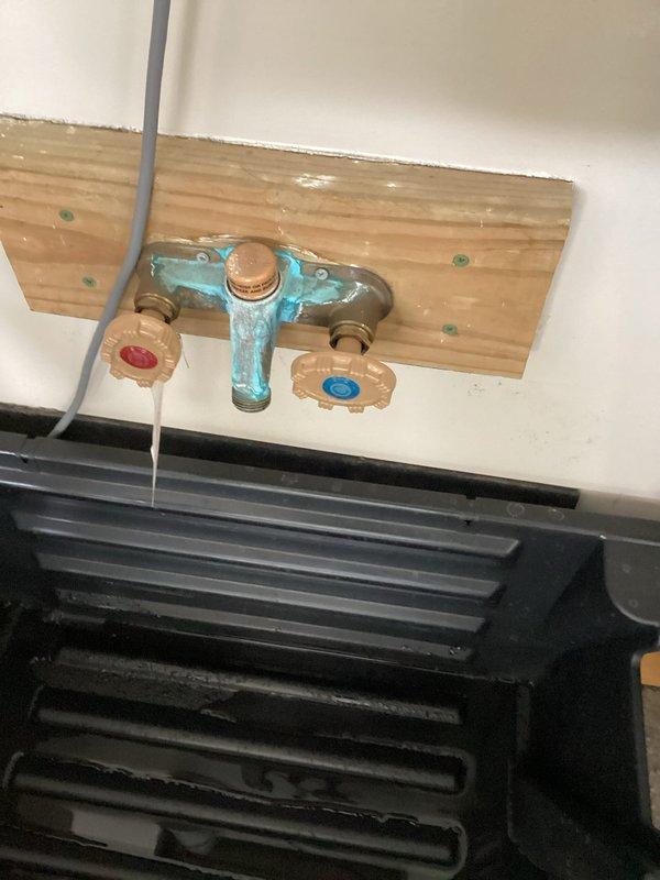 Responded to a plumbing service call for two continuously dripping hose bibbs in a garage office area. Upon inspection, found a dual hot and cold hose bibb assembly mounted on a wooden board showing significant corrosion and constant leaking from both valves. Documented the condition of the turquoise-colored T-shaped brass fixture with photographic evidence and recommended complete replacement of the combined hot and cold water hose bibb assembly to resolve the persistent dripping issue.