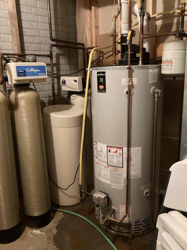 Responded to service call for leaking water heater. Upon arrival, inspected tanked water heater and identified active leak originating from the tank itself. Unit was still producing hot water at time of inspection. Documented equipment condition with photographs showing water heater installation in basement mechanical room alongside water softener system and pressure tank. Assessed leak severity and determined tank failure requiring full water heater replacement. Provided replacement estimate to customer for approval.