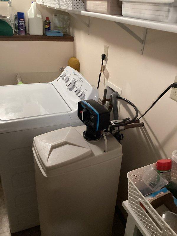Completed installation of water softener system in residential laundry room. Unit was mounted and positioned on top of washing machine, connected to existing plumbing lines, and powered through nearby electrical outlet. System was tested and verified operational upon completion.