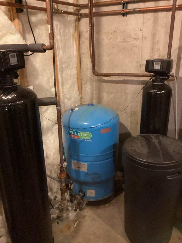Arrived on-site to assess existing water treatment system consisting of an in-place iron filter and water softener. Conducted thorough evaluation of current equipment including a tall black filtration tank with digital control head, blue Well X1000 pressure tank, and existing water softener with associated copper piping and connections. Documented system configuration showing multiple cylindrical tanks with control units, copper supply lines with various fittings and elbows, and electrical wiring connections. Observed moisture staining on concrete floor near equipment base indicating potential system inefficiency. Photographed complete installation including pipe routing, tank positioning, insulated sections, valve components, and spray foam insulation on foundation walls. Prepared comprehensive estimate for replacement iron filter and water softener system to address water quality concerns and improve filtration performance.