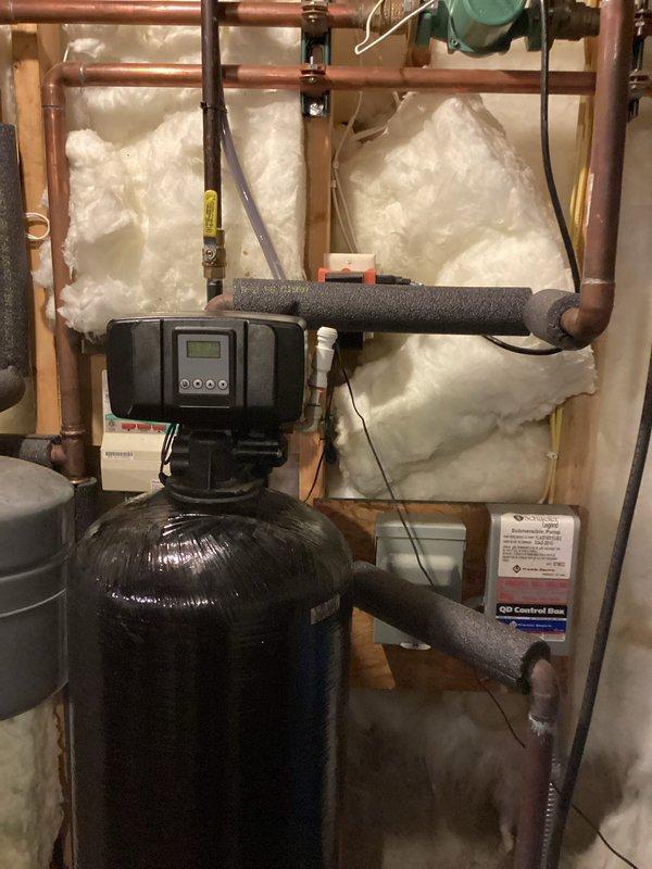 Arrived on-site to assess existing water treatment system consisting of an in-place iron filter and water softener. Conducted thorough evaluation of current equipment including a tall black filtration tank with digital control head, blue Well X1000 pressure tank, and existing water softener with associated copper piping and connections. Documented system configuration showing multiple cylindrical tanks with control units, copper supply lines with various fittings and elbows, and electrical wiring connections. Observed moisture staining on concrete floor near equipment base indicating potential system inefficiency. Photographed complete installation including pipe routing, tank positioning, insulated sections, valve components, and spray foam insulation on foundation walls. Prepared comprehensive estimate for replacement iron filter and water softener system to address water quality concerns and improve filtration performance.