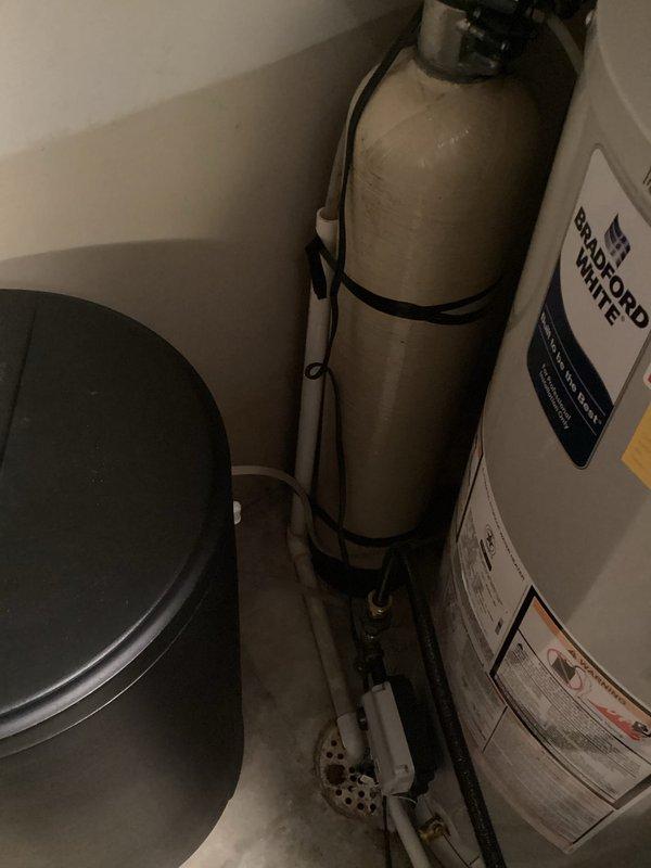 Arrived on site at 10:02 AM to provide water softener installation estimate and complete installation in utility closet area. Assessed existing plumbing configuration including Bradford White water heater, propane cylinder, and available space constraints in confined utility area with slanted ceiling. Provided detailed estimate for water softener system installation, accounting for necessary piping modifications, drain connections, and electrical requirements. Upon approval, proceeded with complete installation of new water softener system with black cylindrical brine tank, securing unit to floor with appropriate strapping per seismic requirements. Connected inlet and outlet water lines using copper piping, installed bypass valve assembly for system maintenance access, and established proper drain line routing. Configured brine tank with salt storage capacity and connected fill line from softener control head. Programmed control valve settings based on water hardness levels and househo