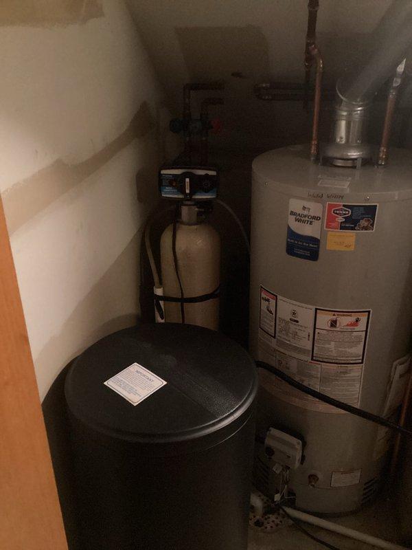 Arrived on site at 10:02 AM to provide water softener installation estimate and complete installation in utility closet area. Assessed existing plumbing configuration including Bradford White water heater, propane cylinder, and available space constraints in confined utility area with slanted ceiling. Provided detailed estimate for water softener system installation, accounting for necessary piping modifications, drain connections, and electrical requirements. Upon approval, proceeded with complete installation of new water softener system with black cylindrical brine tank, securing unit to floor with appropriate strapping per seismic requirements. Connected inlet and outlet water lines using copper piping, installed bypass valve assembly for system maintenance access, and established proper drain line routing. Configured brine tank with salt storage capacity and connected fill line from softener control head. Programmed control valve settings based on water hardness levels and househo