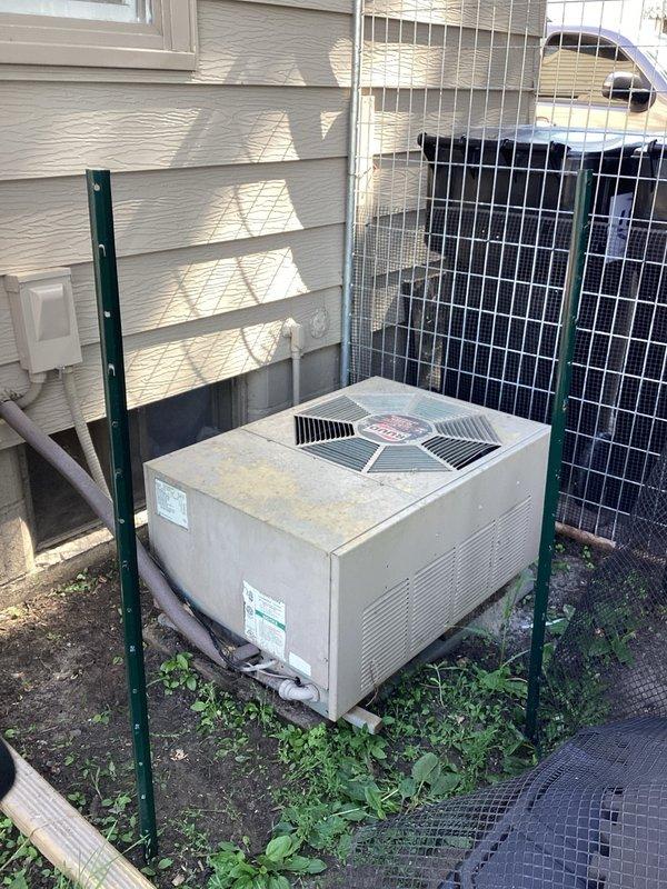 # HVAC Service Call - No Cooling Condition

Responded to a no cooling complaint on a residential air conditioning system. Customer works night shift and scheduled an early morning arrival window. Upon arrival, performed initial system assessment of the outdoor condensing unit, which appeared to be 10+ years old based on visible weathering and condition.

Conducted diagnostic testing and determined the condenser fan motor had failed, preventing proper heat rejection from the refrigeration cycle. The outdoor unit showed signs of age with surface discoloration, dirt accumulation, and weathering consistent with extended outdoor exposure. Unit was positioned on wooden supports near the building exterior with limited clearance to surrounding vegetation.

Documented equipment condition through photographs showing the beige/tan colored condensing unit with its distinctive circular fan grille and manufacturer branding. Visual inspection revealed the unit's location in a side yard area with mini