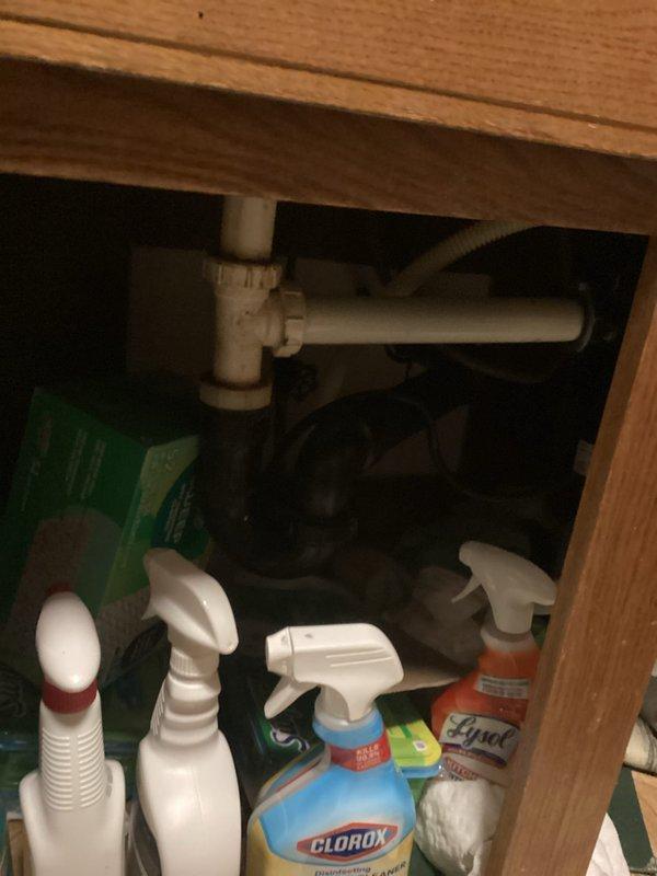 Responded to a service call for a reported leak between the kitchen sink and dishwasher at a rental property. Upon arrival, inspected the under-sink plumbing and cabinet area, examining the white PVC drain assembly including the P-trap configuration and connections. Investigated the reported leak concern and assessed the drain system for potential clogs affecting proper water flow. Documented the existing plumbing configuration and cabinet conditions with photographs showing the sink drain assembly, storage area, and overall kitchen sink installation with stainless steel double-basin sink and fixtures.