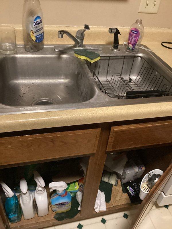Responded to a service call for a reported leak between the kitchen sink and dishwasher at a rental property. Upon arrival, inspected the under-sink plumbing and cabinet area, examining the white PVC drain assembly including the P-trap configuration and connections. Investigated the reported leak concern and assessed the drain system for potential clogs affecting proper water flow. Documented the existing plumbing configuration and cabinet conditions with photographs showing the sink drain assembly, storage area, and overall kitchen sink installation with stainless steel double-basin sink and fixtures.