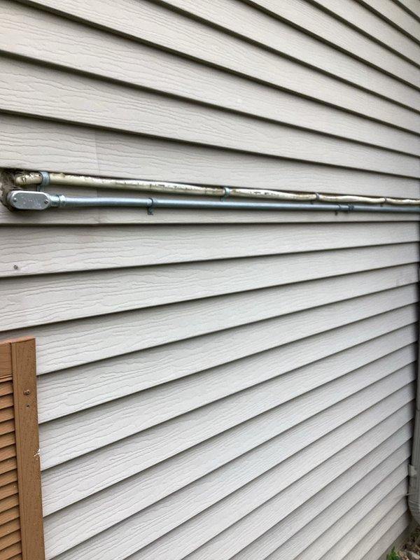Responded to gas leak concern for fireplace insert with exterior gas line installation. Upon inspection, found flexible gas piping (CSST) with severely deteriorated yellow protective jacket exposing bare piping to potential corrosion and weather damage. The exterior-mounted gas line runs along building siding to supply interior brick fireplace with glass doors. Documented existing conditions with photographs showing compromised protective covering on exterior gas line routing. Provided estimate for gas line replacement with recommendation to relocate piping to interior installation for improved protection and longevity.