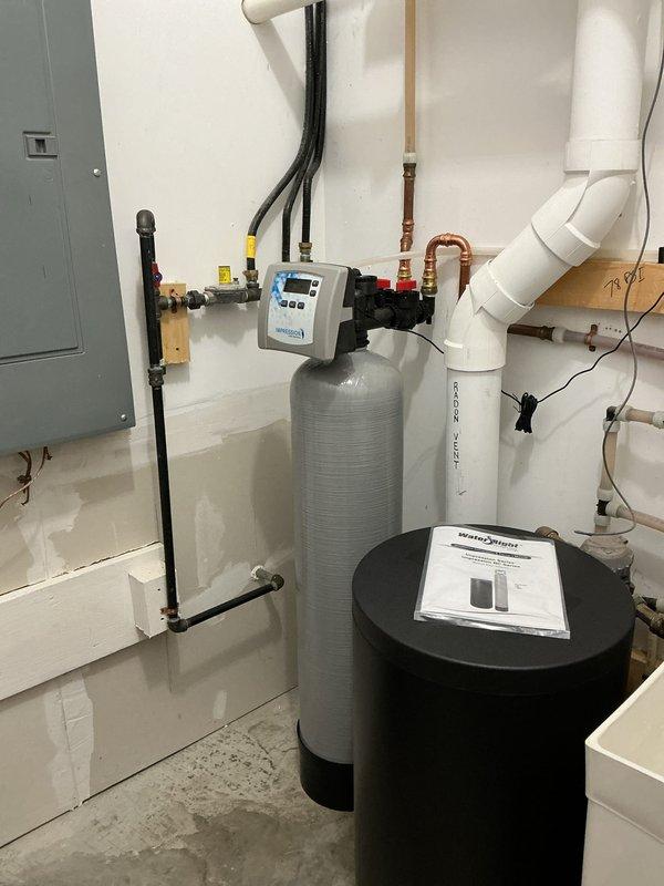 Completed installation of a two-piece Water Right Impressions water softener system in utility room. System includes gray cylindrical softener tank with digital control head, black brine tank, and associated plumbing connections with brass fittings and shutoff valves. All piping connections properly installed and integrated with existing plumbing infrastructure, including copper supply lines and PVC drainage systems. Installation tested and operational.