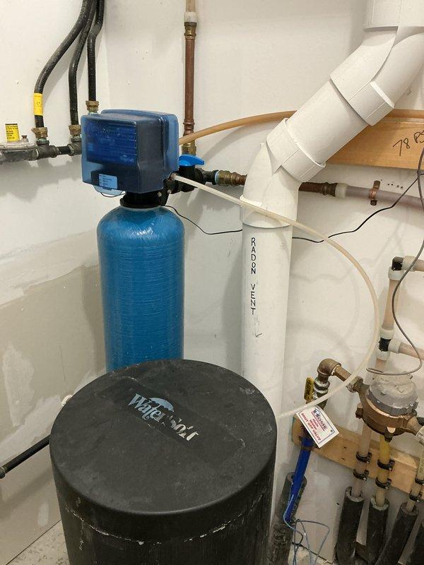 Completed installation of a two-piece Water Right Impressions water softener system in utility room. System includes gray cylindrical softener tank with digital control head, black brine tank, and associated plumbing connections with brass fittings and shutoff valves. All piping connections properly installed and integrated with existing plumbing infrastructure, including copper supply lines and PVC drainage systems. Installation tested and operational.