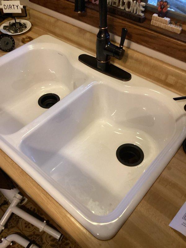 Replaced defective J-trap PVC assembly under white double-basin kitchen sink and installed new drain assemblies for both basins. All plumbing connections were secured and tested for proper drainage, with white PVC piping configured to accommodate the dual-basin setup with black drain fittings.