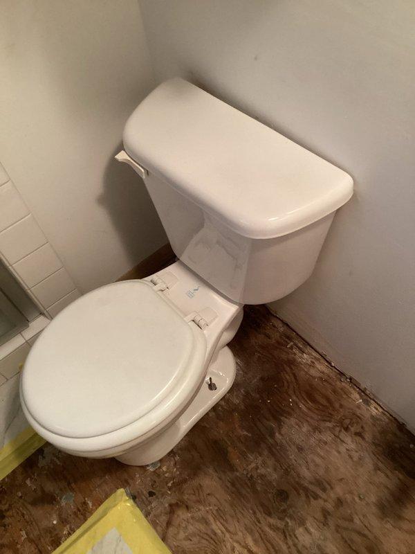 Reinstalled white two-piece toilet in master bathroom corner following demolition work. Toilet fixture secured and positioned against subway-tiled wall with proper mounting and seal. Significant pre-existing water damage observed on surrounding flooring, displaying extensive dark staining and deterioration around toilet base, indicating potential long-term moisture exposure requiring future attention.