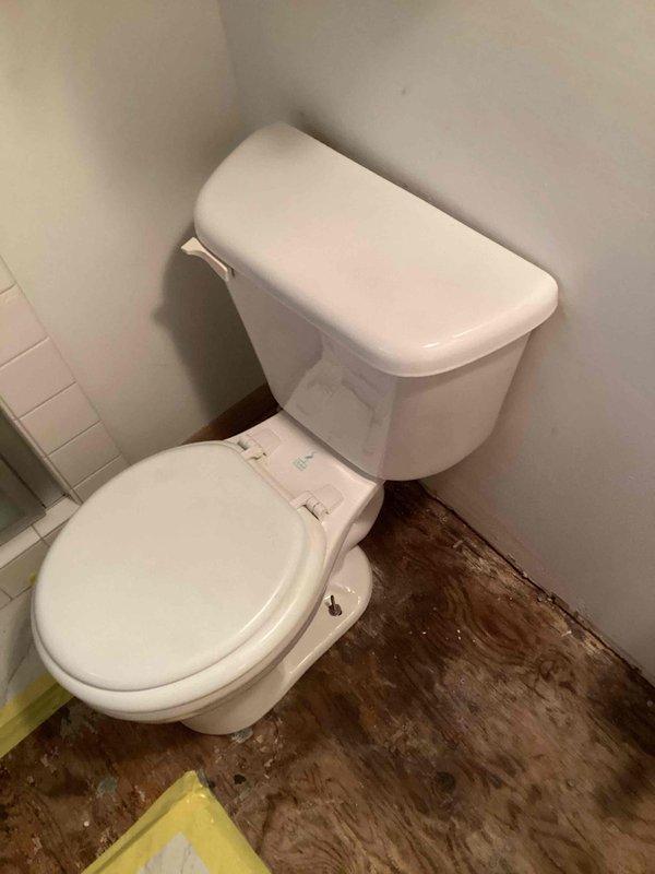 Reinstalled white two-piece toilet in master bathroom corner following demolition work. Toilet fixture secured and positioned against subway-tiled wall with proper mounting and seal. Significant pre-existing water damage observed on surrounding flooring, displaying extensive dark staining and deterioration around toilet base, indicating potential long-term moisture exposure requiring future attention.