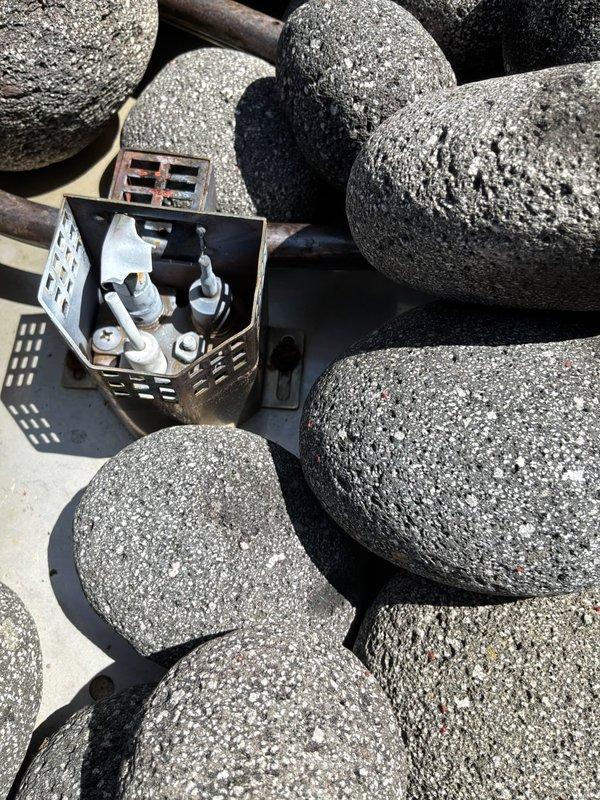 Responded to service call for non-operational outdoor fireplace located in screened porch area. Upon arrival, inspected wood-burning stove installation and surrounding components. Examined gas fire pit assembly with decorative lava rock arrangement and identified issues with burner system components including damaged or improperly configured gas supply fittings visible in metal basket assembly. Assessed venting system, stovepipe installation, and clearances to combustible materials. Documented existing conditions with photographs for follow-up service requirements.