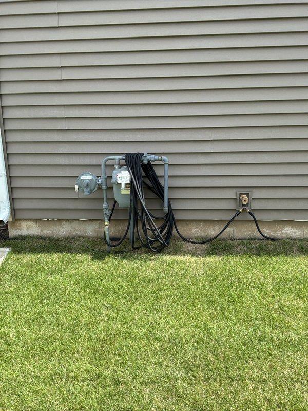Windsor, WI - Conducted on-site evaluation for whole house standby generator installation at residential property. Assessed existing electrical service panel located in unfinished basement area and exterior utility infrastructure including natural gas meter and service entrance cabling. Discussed power requirements with homeowner to determine appropriate generator sizing for complete home coverage during utility outages. Evaluated two generator options: 18kW unit and 23/26kW unit, with consideration for air conditioning load requirements and potential soft start integration. Identified need for natural gas meter upsizing and installation of gas pressure regulator to accommodate generator fuel demand in addition to existing gas appliances. Documented current electrical panel configuration and available installation space on exterior wall adjacent to existing gas meter location. Photographed utility area showing gas meter, electrical service entrance cables, and outdoor receptacle for equipment placem