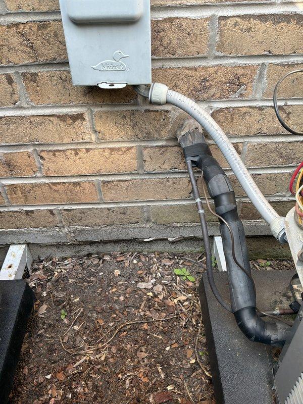 Responded to service call for non-operational Amana condenser unit (Model ASX130301) and recurring NTI boiler leak. Upon arrival, inspected outdoor electrical disconnect and conduit system serving the 11-year-old AC unit. Found electrical disconnect box mounted on exterior brick wall with visible weathering and debris accumulation at base. Verified equipment specifications and confirmed condenser was not receiving power. Inspected flexible conduit connections and weatherproof fittings for proper continuity. Documented electrical service installation and exterior mounting conditions with photos showing disconnect enclosure, conduit routing, and connection points. Addressed power supply issue to restore cooling system operation and evaluated boiler leak condition for necessary repairs.