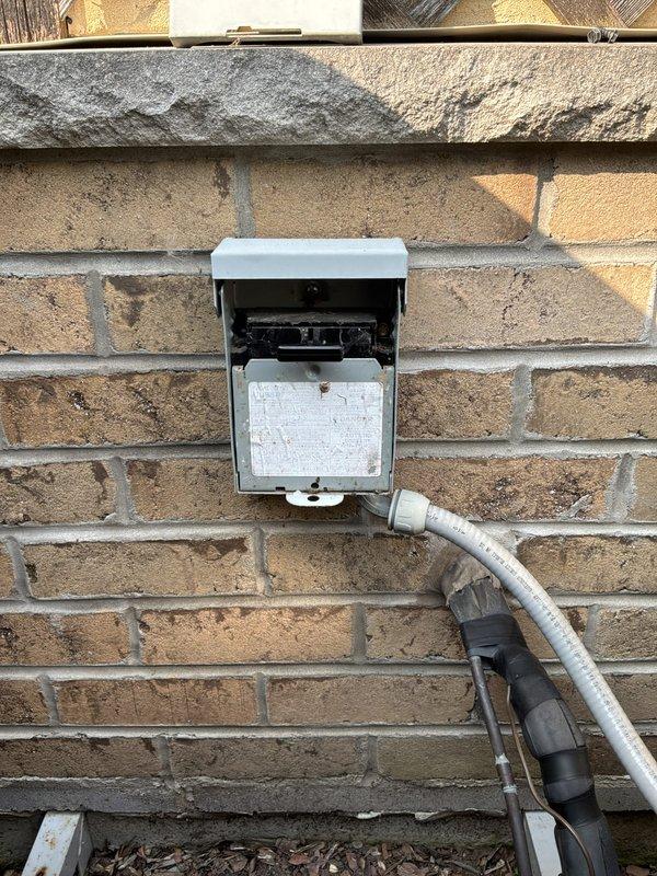 Responded to service call for non-operational Amana condenser unit (Model ASX130301) and recurring NTI boiler leak. Upon arrival, inspected outdoor electrical disconnect and conduit system serving the 11-year-old AC unit. Found electrical disconnect box mounted on exterior brick wall with visible weathering and debris accumulation at base. Verified equipment specifications and confirmed condenser was not receiving power. Inspected flexible conduit connections and weatherproof fittings for proper continuity. Documented electrical service installation and exterior mounting conditions with photos showing disconnect enclosure, conduit routing, and connection points. Addressed power supply issue to restore cooling system operation and evaluated boiler leak condition for necessary repairs.