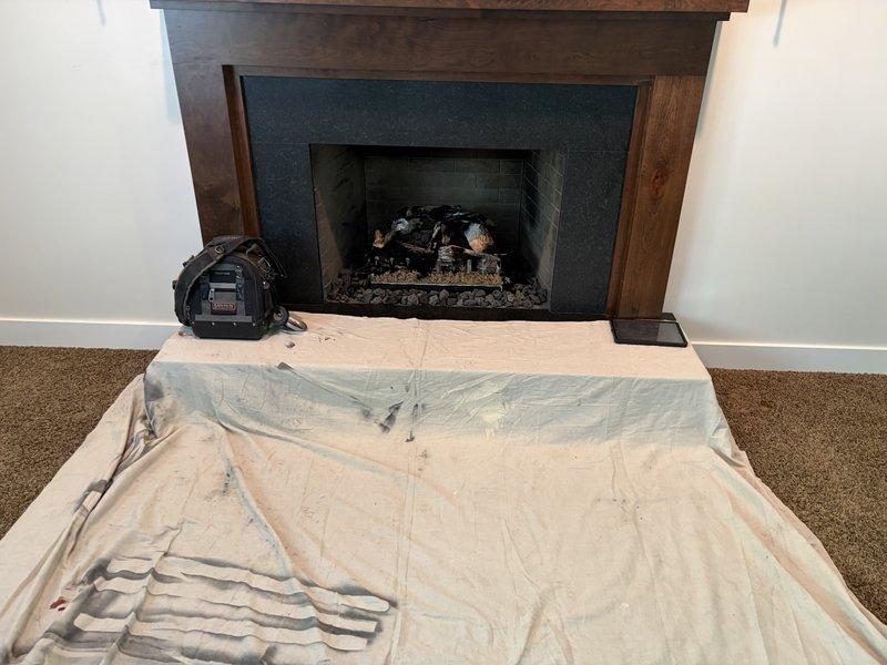 Returned to property to complete fireplace makeover installation. Replaced existing fireplace surround with new dark teal/blue-gray surround material and reinstalled wooden mantel framing. Verified proper operation of gas fireplace unit with artificial log set and decorative rock bed. Tested ignition system and confirmed clean flame pattern with appropriate yellow and orange coloration. Protected work area with drop cloths throughout installation process. All components functioning properly upon completion.