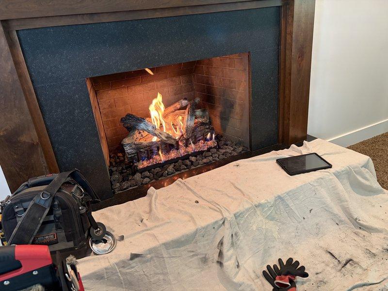 Returned to property to complete fireplace makeover installation. Replaced existing fireplace surround with new dark teal/blue-gray surround material and reinstalled wooden mantel framing. Verified proper operation of gas fireplace unit with artificial log set and decorative rock bed. Tested ignition system and confirmed clean flame pattern with appropriate yellow and orange coloration. Protected work area with drop cloths throughout installation process. All components functioning properly upon completion.
