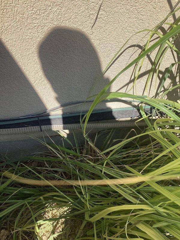 Responded to service call for 8-year-old Coleman AC system blowing warm air with inadequate cooling performance. Upon arrival, confirmed unit was not iced over as customer had suspected. Conducted comprehensive system inspection of outdoor condensing unit located behind south gate and indoor air handler components. Outdoor unit found with overgrown vegetation obstructing airflow around condenser coil, which can significantly impact heat rejection and system efficiency. Indoor inspection revealed heavily corroded copper refrigerant lines and deteriorated piping throughout air handler area, indicating long-term moisture exposure and lack of preventive maintenance. Corrosion observed on multiple pipe sections poses risk for future refrigerant leaks and system failure. Electrical connections and wiring inspected with no immediate safety concerns noted. System operational at time of inspection but operating with compromised efficiency due to restricted airflow and component deterioration. D