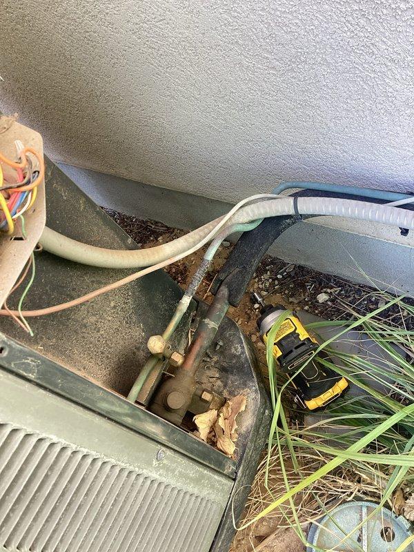 Responded to service call for 8-year-old Coleman AC system blowing warm air with inadequate cooling performance. Upon arrival, confirmed unit was not iced over as customer had suspected. Conducted comprehensive system inspection of outdoor condensing unit located behind south gate and indoor air handler components. Outdoor unit found with overgrown vegetation obstructing airflow around condenser coil, which can significantly impact heat rejection and system efficiency. Indoor inspection revealed heavily corroded copper refrigerant lines and deteriorated piping throughout air handler area, indicating long-term moisture exposure and lack of preventive maintenance. Corrosion observed on multiple pipe sections poses risk for future refrigerant leaks and system failure. Electrical connections and wiring inspected with no immediate safety concerns noted. System operational at time of inspection but operating with compromised efficiency due to restricted airflow and component deterioration. D