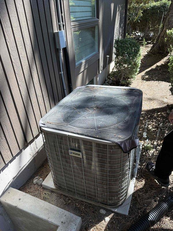 Responded to service call for York air conditioning condenser unit, approximately 15-20 years old, with reported failure to cool and inoperative condenser fan. Upon arrival, performed visual inspection of outdoor condenser unit positioned on concrete pad adjacent to building exterior. Unit showed signs of age and weathering with dust accumulation on protective cover. Conducted diagnostic evaluation to assess system performance and identify root cause of cooling failure and fan malfunction. Homeowner also reported unusual chemical odor present in system operation. Documented equipment condition through photography showing condenser placement, electrical disconnect, and overall installation configuration. Provided technical assessment and repair recommendations based on findings from diagnostic evaluation.