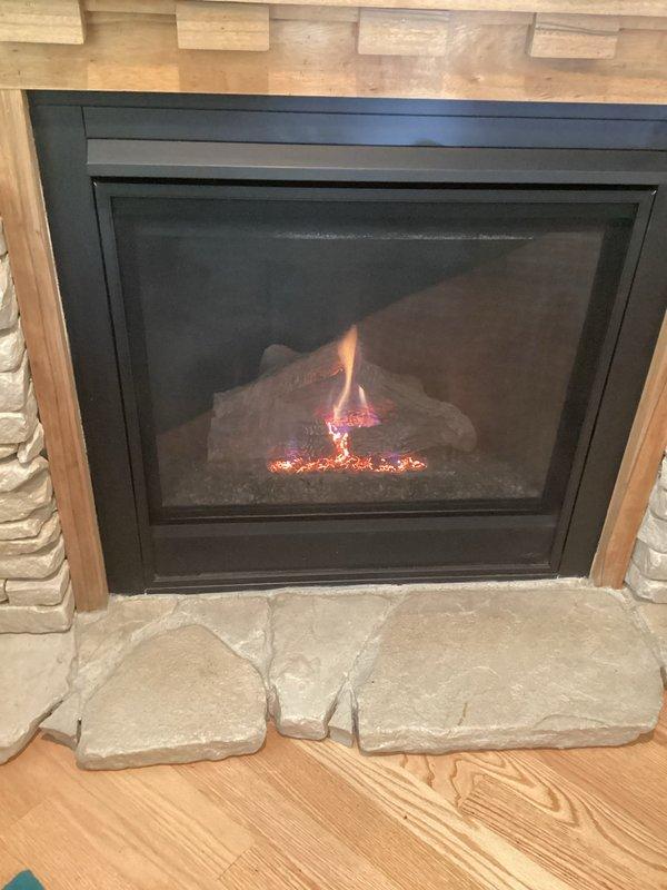 Responded to service call for 16-year-old gas fireplace experiencing intermittent cycling issues, with unit running continuously at times or shutting off after approximately 30 minutes before restarting. Upon inspection, observed significant corrosion and deterioration on burner assembly and heat exchanger components, which can cause improper flame sensing and safety system malfunctions leading to erratic operation. Documented current condition of gas fireplace insert with stone surround in living room, noting the corroded burner surfaces and accumulated debris in combustion chamber that likely contribute to inconsistent flame detection and automatic shutdown cycles.