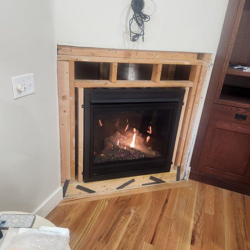 Completed installation of gas fireplace unit with black frame and glass front insert. Framed opening with dimensional lumber, installed flexible ductwork for proper ventilation, and terminated vent pipe through roof with appropriate flashing and weather boot. Fireplace unit tested and operational with visible flame pattern and decorative log set. Electrical connections completed with wall-mounted controls installed adjacent to unit.