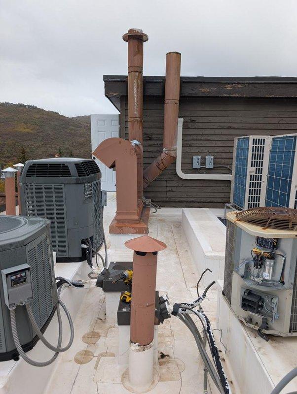Technicians conducted a comprehensive site assessment of the rooftop HVAC system to evaluate conditions and requirements for an upcoming installation. Detailed photographic documentation was completed of all existing equipment, including multiple air conditioning units with protective caging, copper ventilation piping with weathered patina, exhaust stacks with rain hoods, electrical components, and adjacent solar panel arrays to ensure accurate planning for the future work.