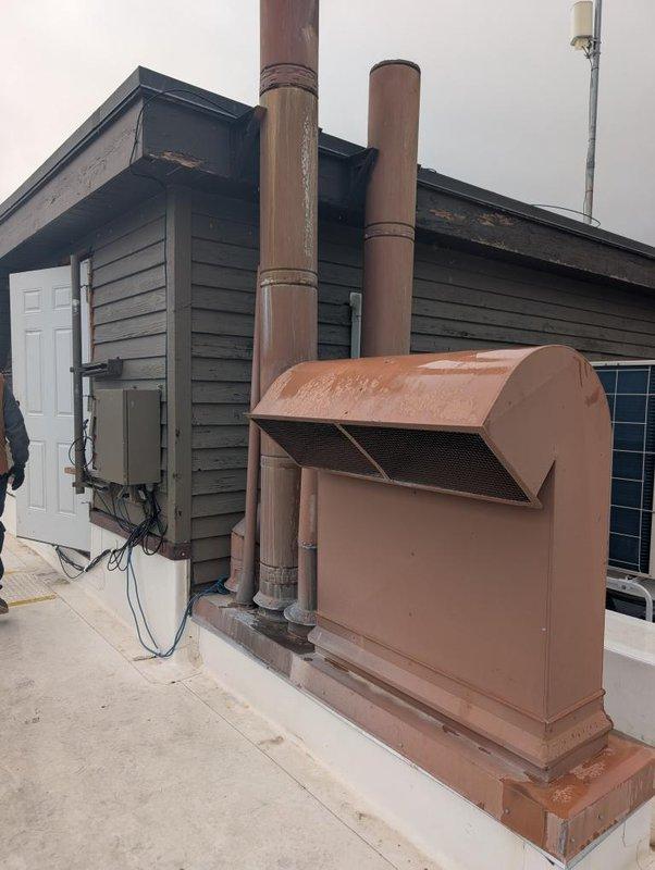 Technicians conducted a comprehensive site assessment of the rooftop HVAC system to evaluate conditions and requirements for an upcoming installation. Detailed photographic documentation was completed of all existing equipment, including multiple air conditioning units with protective caging, copper ventilation piping with weathered patina, exhaust stacks with rain hoods, electrical components, and adjacent solar panel arrays to ensure accurate planning for the future work.