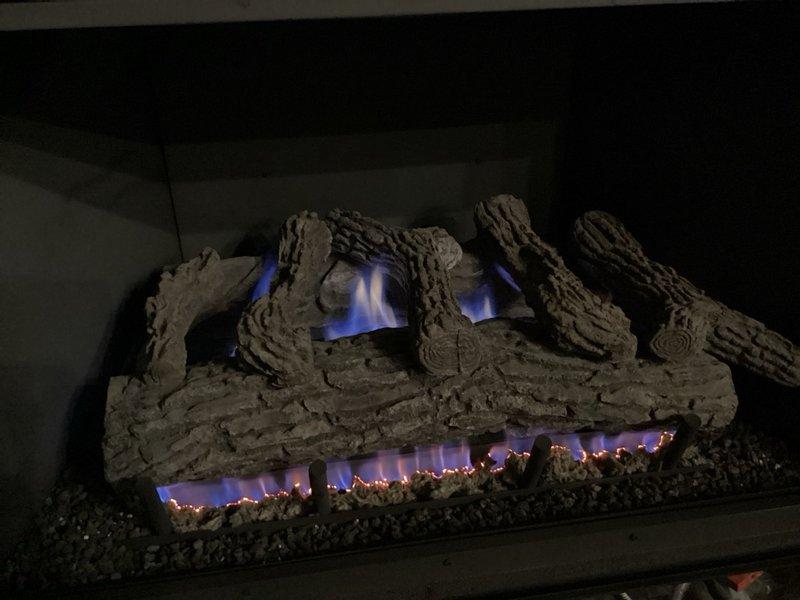 Responded to a callback on a recently upgraded gas fireplace reporting predominantly blue flames with minimal yellow and one burner flame remaining entirely blue. Inspected the firebox, ceramic log set, and burner pan, and found dust, soot, and debris accumulation around the burner ports and base of the burner assembly. Cleaned the burner area and observed the flame pattern after adjustment, noting blue flames with orange and pink tips along the burner and visible ember glow on the log set.