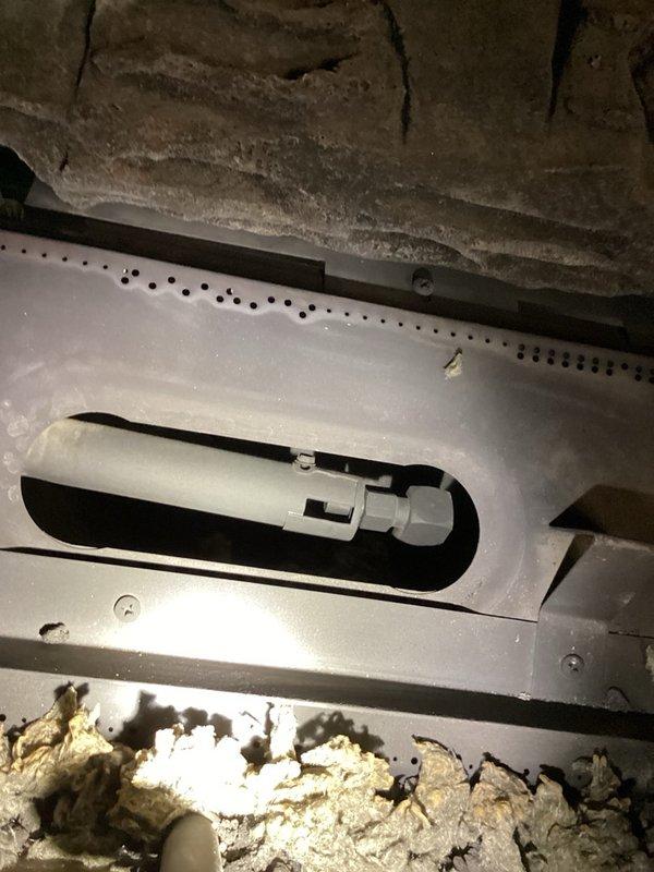 Responded to a callback on a recently upgraded gas fireplace reporting predominantly blue flames with minimal yellow and one burner flame remaining entirely blue. Inspected the firebox, ceramic log set, and burner pan, and found dust, soot, and debris accumulation around the burner ports and base of the burner assembly. Cleaned the burner area and observed the flame pattern after adjustment, noting blue flames with orange and pink tips along the burner and visible ember glow on the log set.