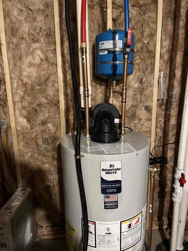 Successfully installed a Bradford White water heater in utility area, complete with all necessary connections including copper pipes with brass fittings and PVC piping. System includes a properly mounted blue expansion tank and appropriate valving, with all components verified to be in good working condition with no leaks or damage.